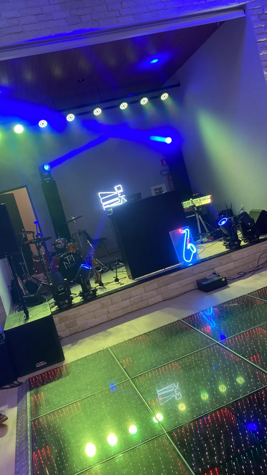 Palco montado no salão, com cabine de DJ, bateria, saxofone e luzes azuis, e pista de LED verde à frente